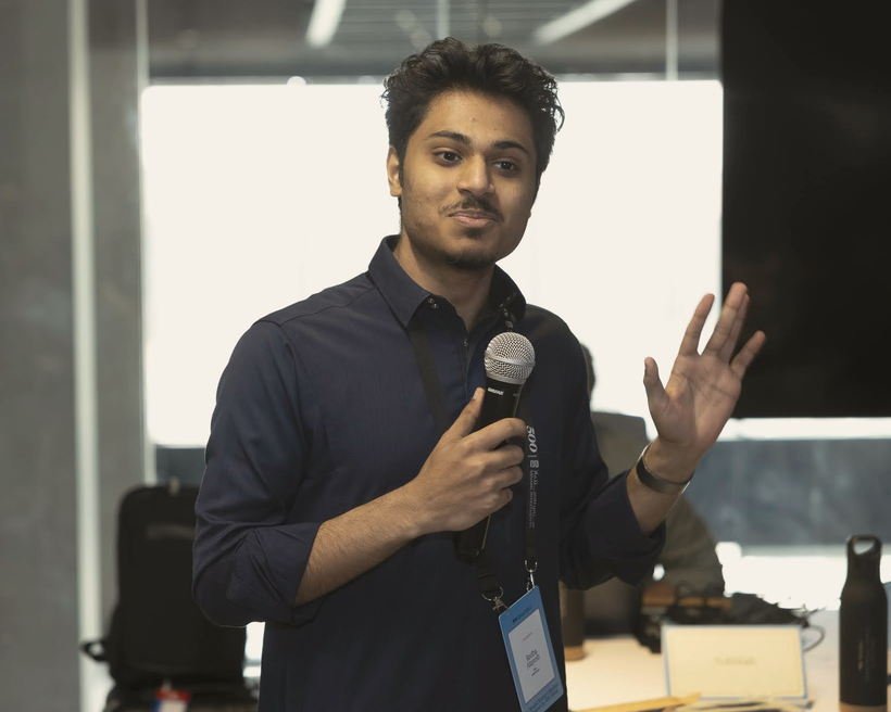 Redha Alayesh speaking to a room with a microphone in one hand, the other hand raised mid-explanation.