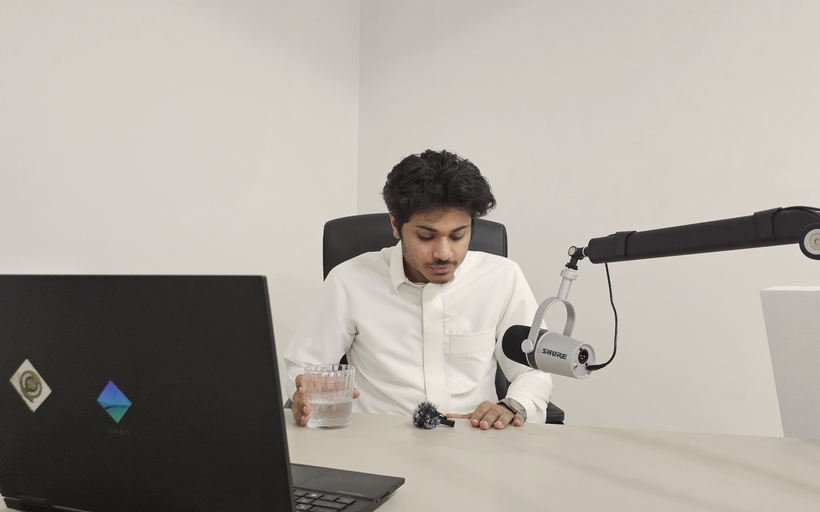 Redha Alayesh at a plain desk with a laptop and a microphone on a boom arm, the room where the Arabic videos are recorded.